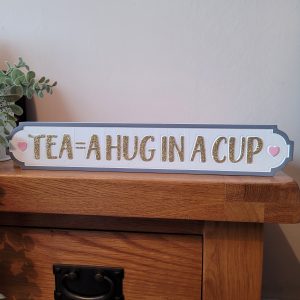 A white free standing block with a dark grey border with gold glitter text 'Tea = A Hug In A Cup'. With cute pink hearts either side, a fabulous sign for tea lovers!