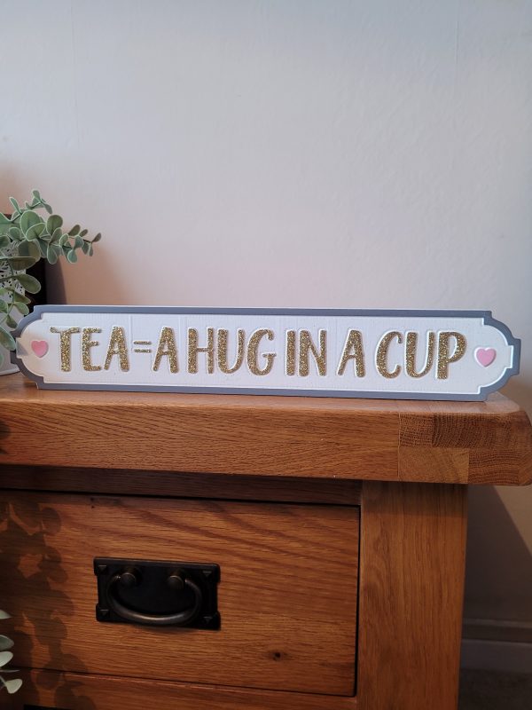 A white free standing block with a dark grey border with gold glitter text 'Tea = A Hug In A Cup'. With cute pink hearts either side, a fabulous sign for tea lovers!